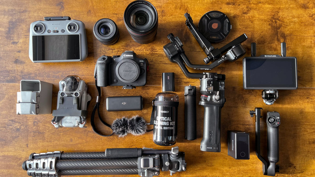 A curated flat-lay collection of professional camera gear and accessories arranged neatly on a warm wooden surface. The collection includes a Mirrorless Canon R6 Mark II camera body, multiple lenses, a gimbal, a monitor screen, a tripod, a Drone and remote controller, a battery, a small fuzzy wind shield, and other mounting accessories.