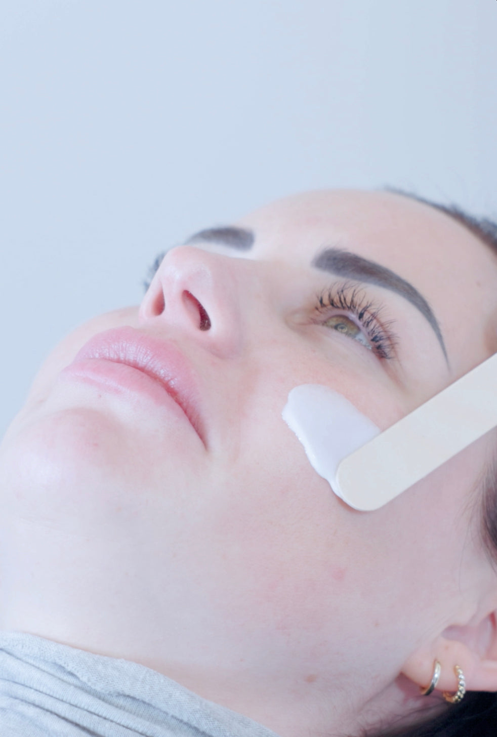 Close-up of a woman lying down with a white cosmetic cream being spread onto her cheek using a spatula, against a light gray background. Photo taken by Photographer Thiago Mazzeo