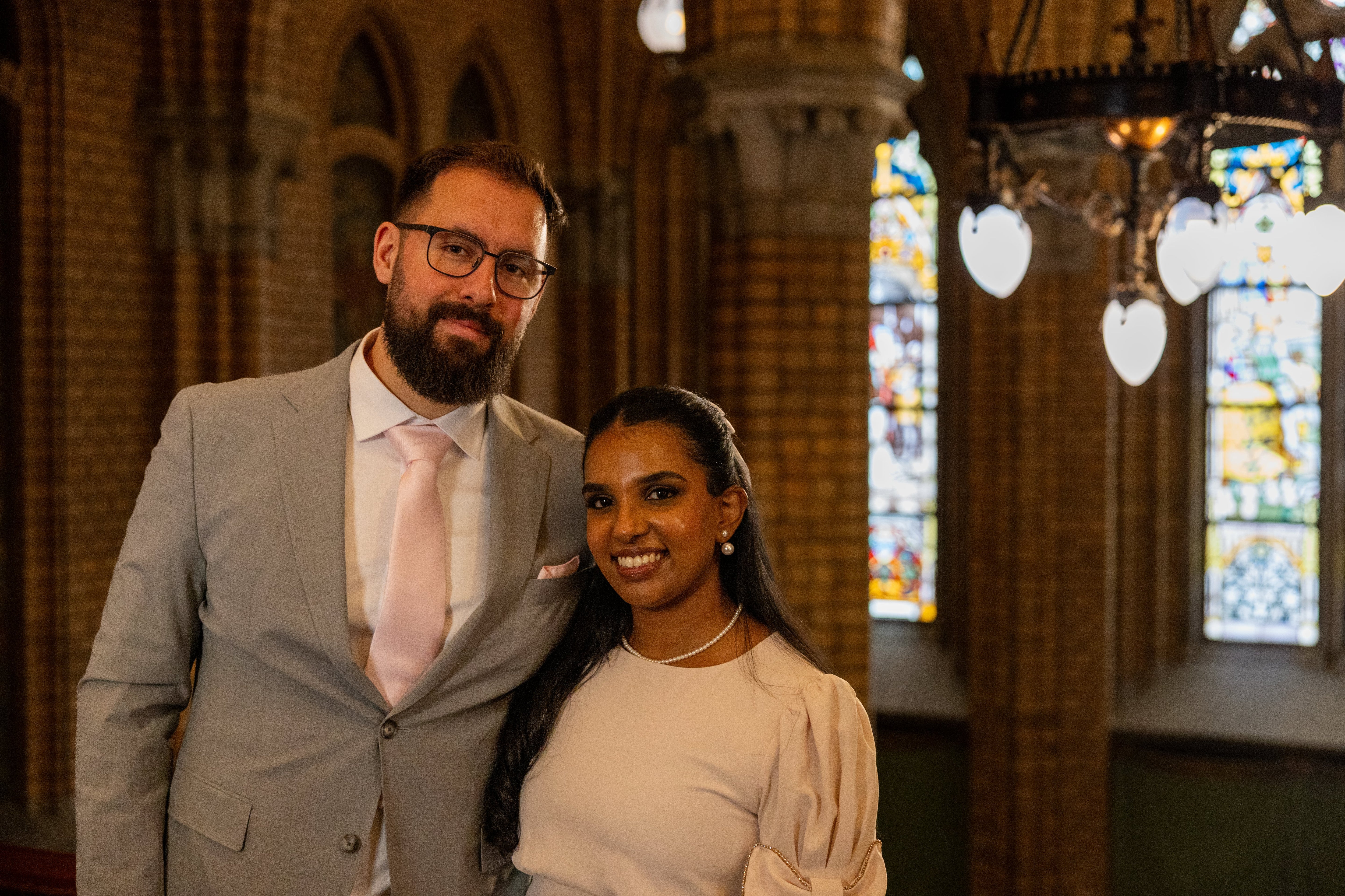 A man in a light gray suit and a woman in a cream puff-sleeve top standing together inside a church with brick columns and stained glass windows. Photo taken by Photographer Thiago Mazzeo