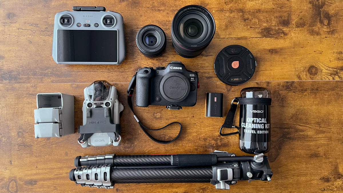 A top-down flat lay of photography equipment arranged on a wooden surface, including a drone, controller, two camera lenses, a Canon R6 Mark II Mirrorless camera body, a lens filter, a drone battery, a tripod, and a cleaning kit.