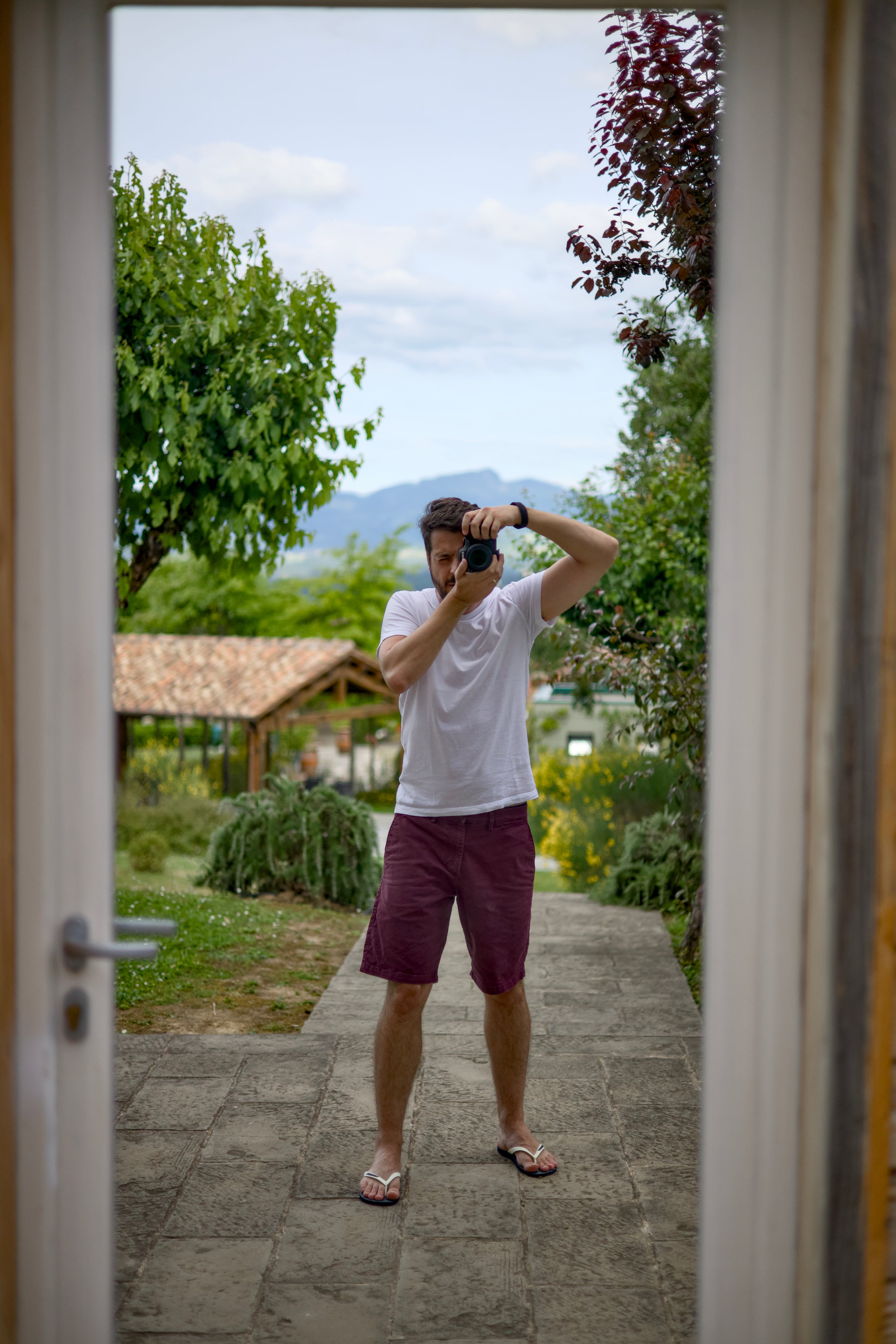 Photographer Thiago Mazzeo wearing a white t-shirt and maroon shorts stands on a stone path holding a camera, framed by an open doorway with a garden and mountains in the background.