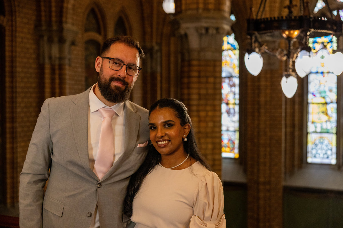 A man in a light gray suit and a woman in a cream puff-sleeve top standing together inside a church with brick columns and stained glass windows. Photo taken by Photographer Thiago Mazzeo