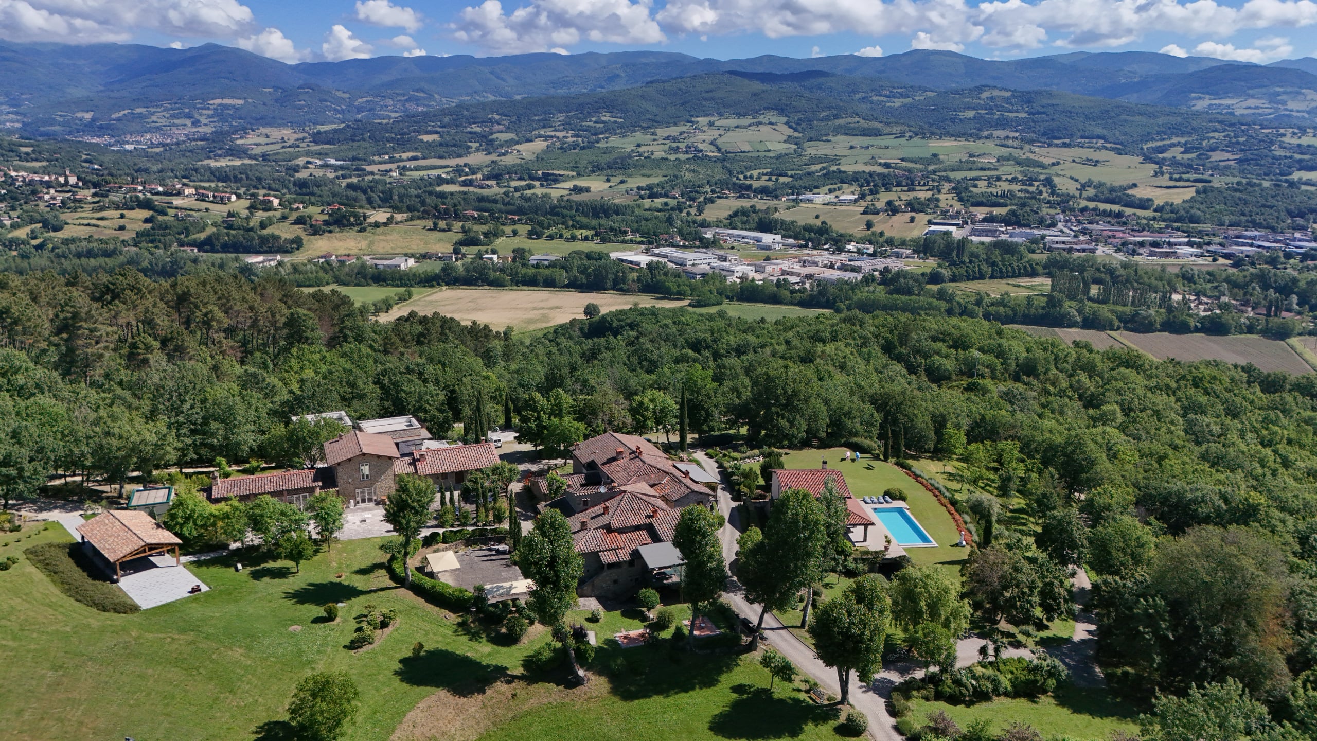 Aerial view of a large estate with multiple buildings, a swimming pool, extensive green lawns and trees, and a mountain valley landscape in the background in Tuscany, Italy.